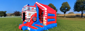 Rote und blaue Hüpfburg gestaltet wie ein Feuerwehrauto mit Rutsche vor grünem Wiesenhintergrund