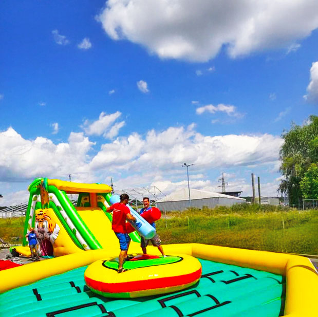 Zwei Erwachsene die gegeneinander in einer Gladiatoren Arena antreten, im Hintergrund steht eine Riesenrutsche
