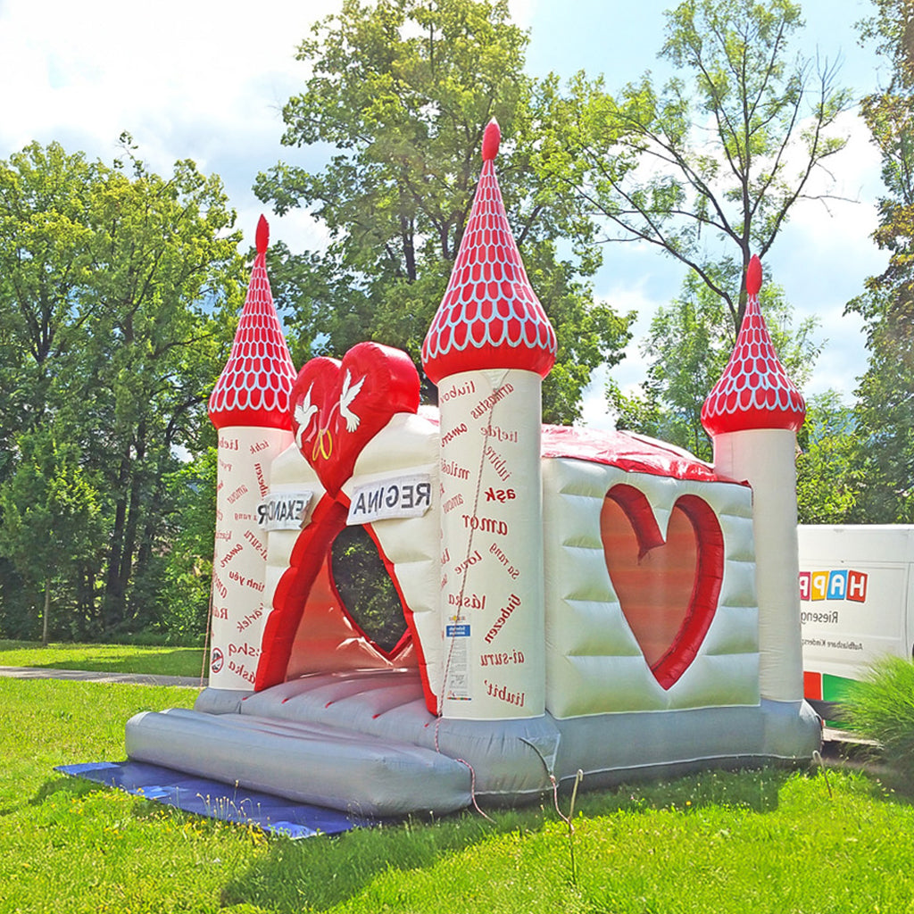 Eine Hüpfburg mit rotem Dach, roten spitzen Schlosstürmen, weißen Wänden mit Herzform Fenstern und graue Hüpffläche.