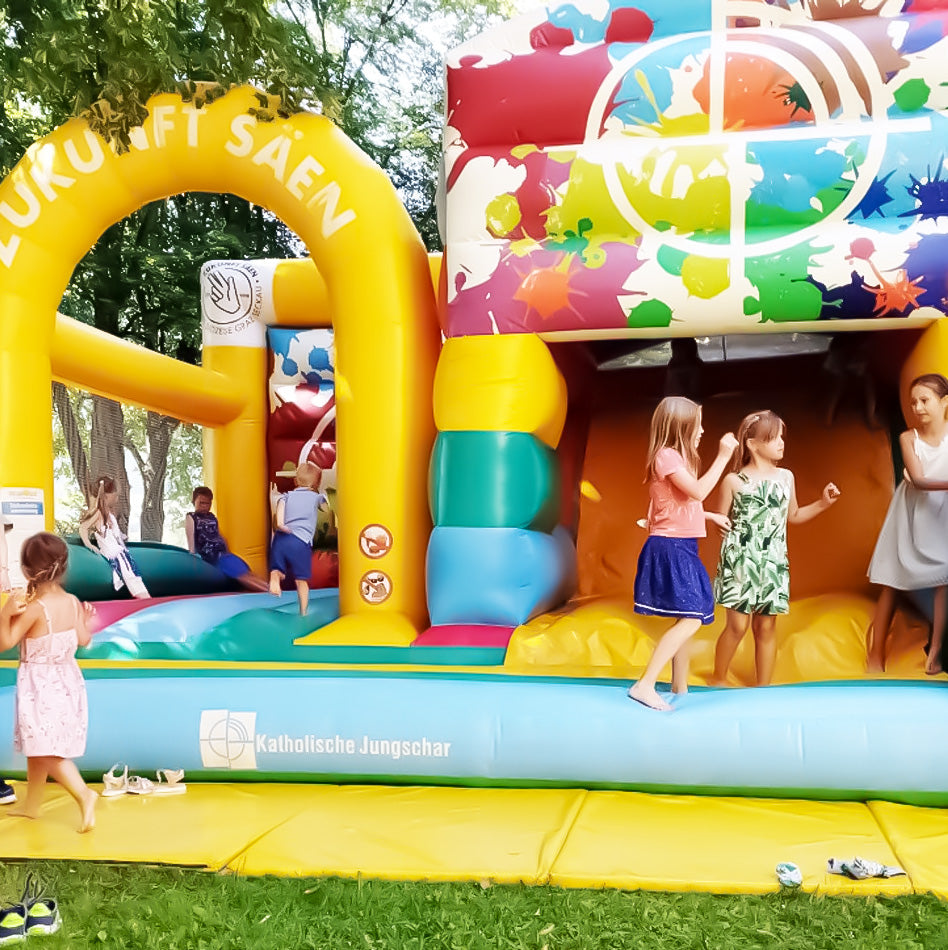 Kinder toben auf bunter Kirchen-Hüpfburg mit dem Slogan „Zukunft säen“ der Katholischen Jungschar im Park