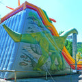 Aufblasbare Dino-Jumprutsche mit zwei Rutschbahnen, Dino-Dekor und Palmen – farbenfrohe Hüpfburg im Jurassic-Design