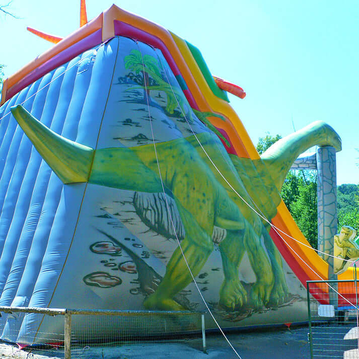 Aufblasbare Dino-Jumprutsche mit zwei Rutschbahnen, Dino-Dekor und Palmen – farbenfrohe Hüpfburg im Jurassic-Design