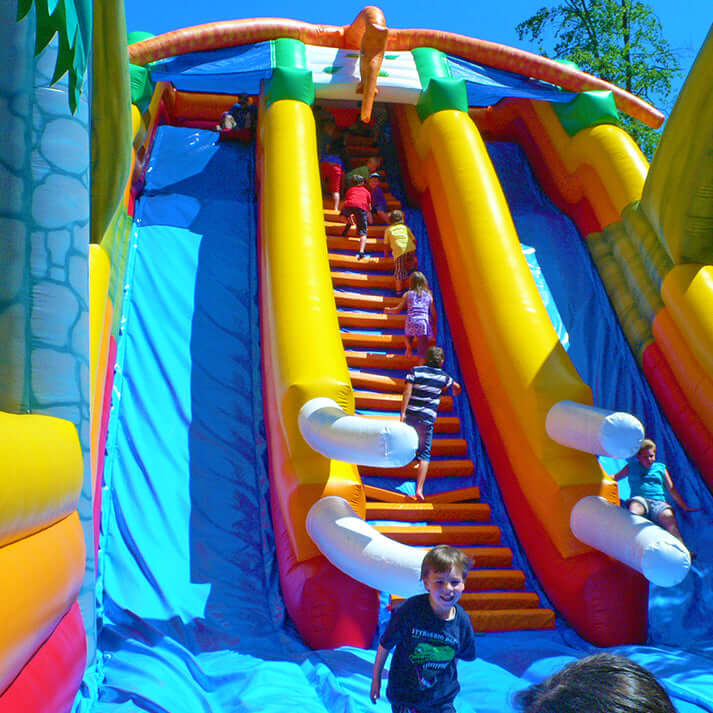 Aufblasbare Dino-Jumprutsche mit zwei Rutschbahnen, Dino-Dekor und Palmen – farbenfrohe Hüpfburg im Jurassic-Design