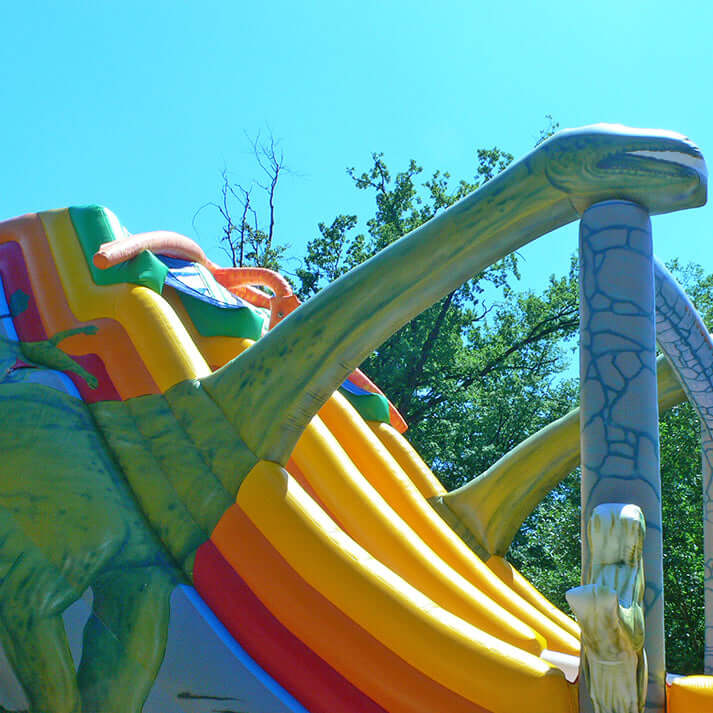 Aufblasbare Dino-Jumprutsche mit zwei Rutschbahnen, Dino-Dekor und Palmen – farbenfrohe Hüpfburg im Jurassic-Design