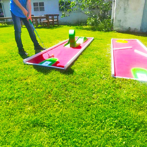 Zwei Minigolfbahnen in rot mit Hindernissen auf einer gruenen Wiese und ein Mann neben der Bahn mit einem Minigolf Schlaeger