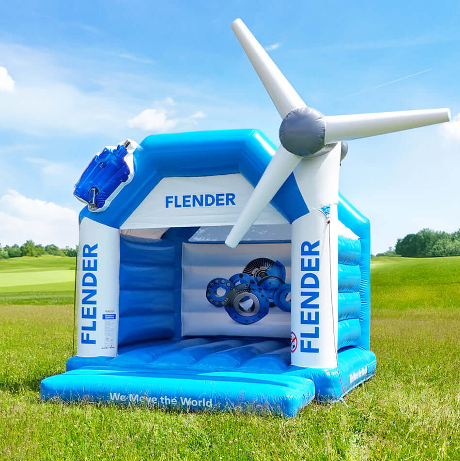 Aufblasbare Hüpfburg in Blau und Weiß mit dem Aufdruck „Flender“, dekoriert mit einem Windrad auf einer grünen Wiese bei Sonnenschein.