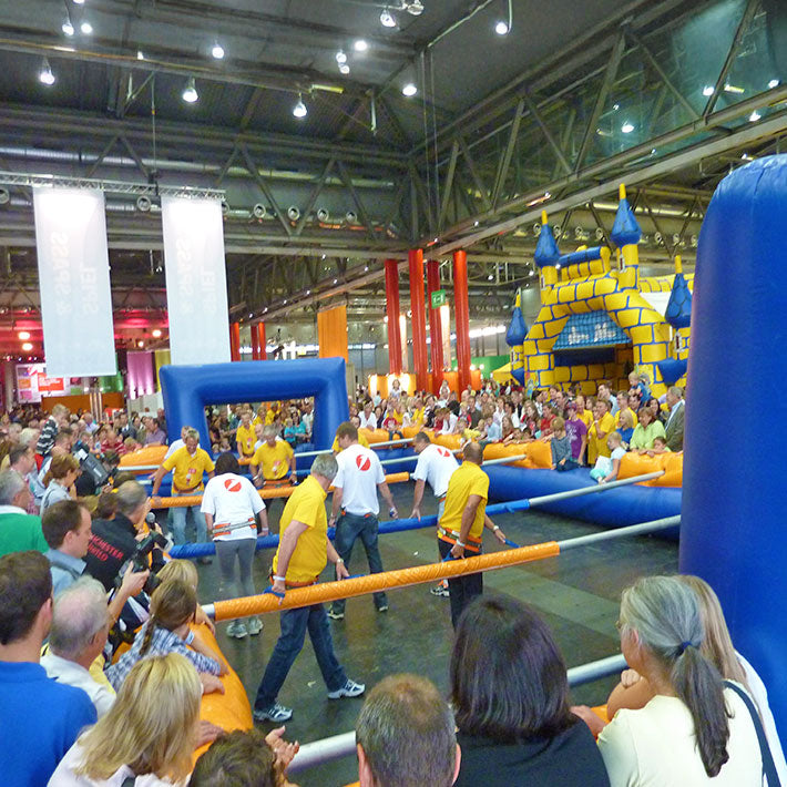 Zahlreiche Menschen die bei einem Riesenwuzzler Turnier zusehen der in einer Halle eben Huepfburgen steht.