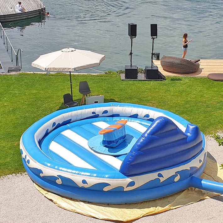 Aufblasbarer Surf-Simulator in Blau-Weiss gehalten neben einem Sonnenschirm bei einem See.