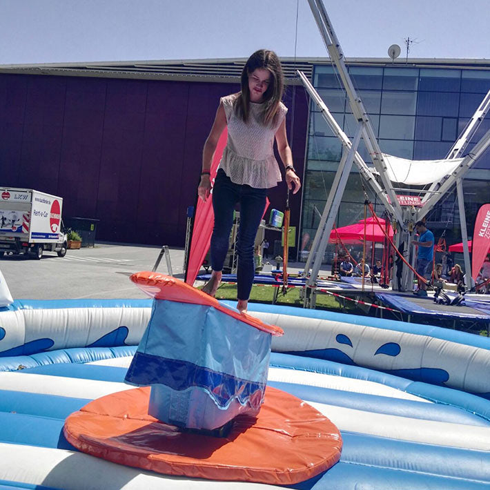 Junges Maedchen auf einem Aufblasbarem Surf-Simulator neben einem Trampolin.