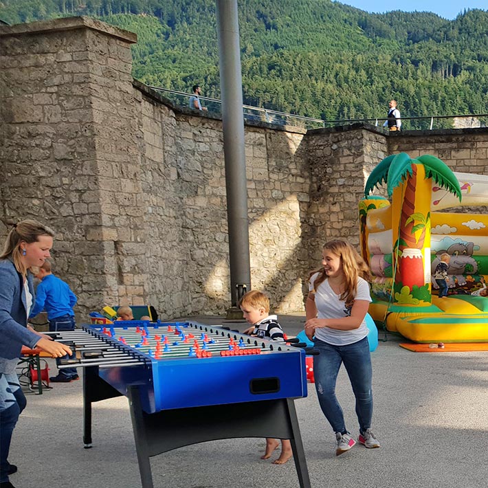 Zwei Frauen und ein Kind die auf einem Tischwuzzler für acht Personen spielen der neben einer aufblasbaren Huepfburg steht.