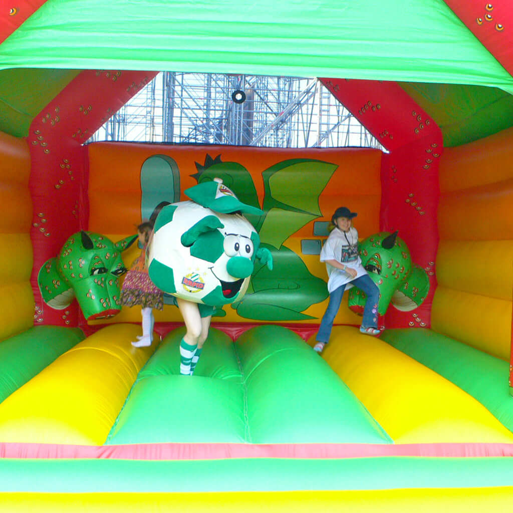 Hüpfburg mit grün gelber Sprungfläche, zwei Elementen als Drachenköpfe und spielenden Kindern.