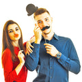 Eine Frau die sich rote Lippen zum Mund haelt und eine Mann der sich einen Hut auf den Kopf haelt und einen Schnurbart ober die Lippe