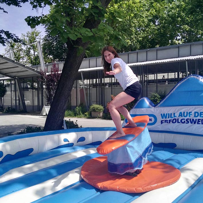 Junge Frau auf einem Surf-Simulator mit Grau-Orangenen Surfbrett im freien.