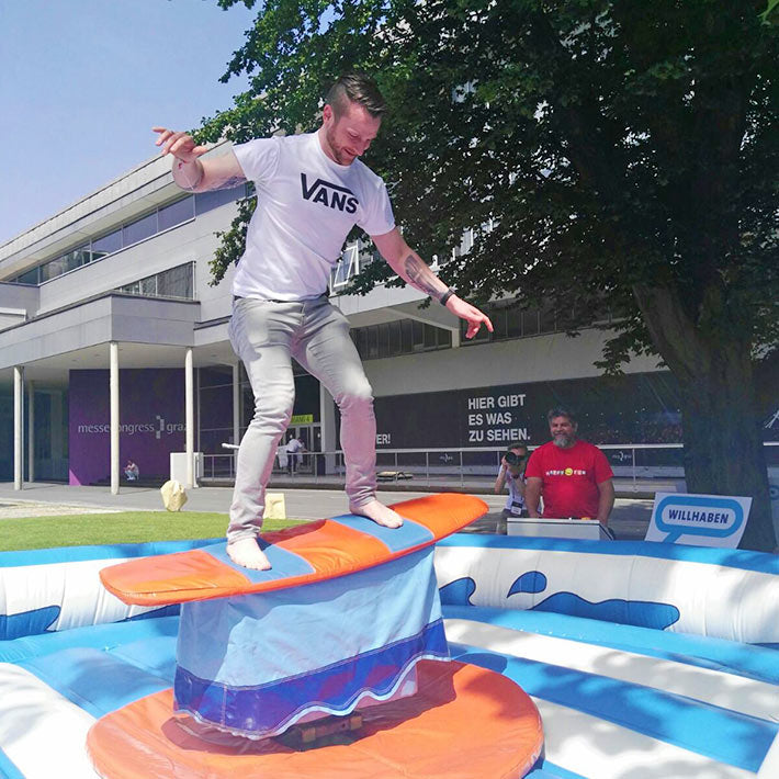 Ein Mann auf einem Surf-Simulator im freien neben einem grossen Baum und ein Mann in einem Roten Shirt, der das Steuerpult bedient. 