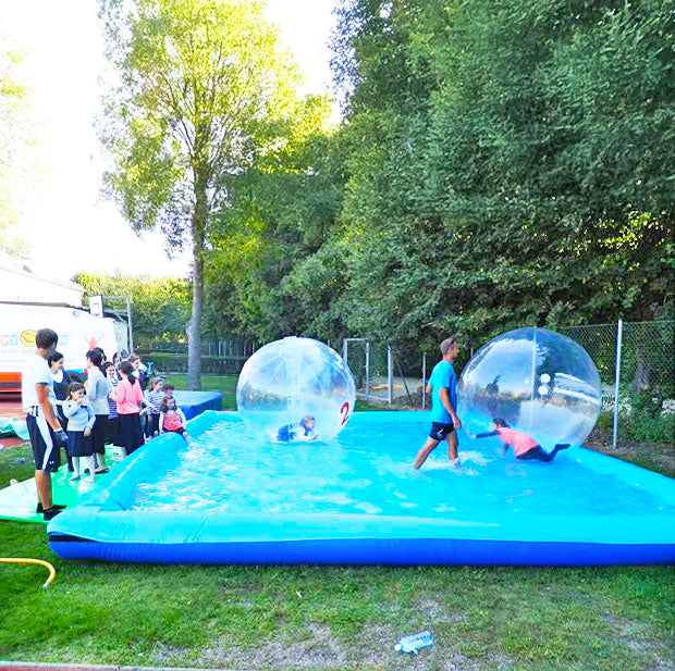 Froehliche Kinder in einem aufblasbarem Wasserbecken in beschrifteten Wasserbaellen und Kinder die am Rand zusehen.