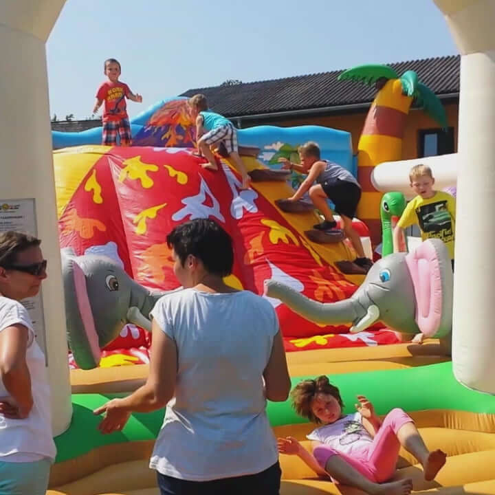 Kinder die einen Vulkan in einer Huepfburg raufklettern und wieder runter rutschen und zwei aufblasbare Elefanten mit langen Ruessel.