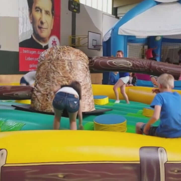 Kindgerechte Hüpfburg Heuhüpfer mit ländlichem Design und großen Heuballen als Gestaltungselemente, Kinder springen ausgelassen in sicherer Umgebung und weichen einem sich drehenden Hindernis aus.