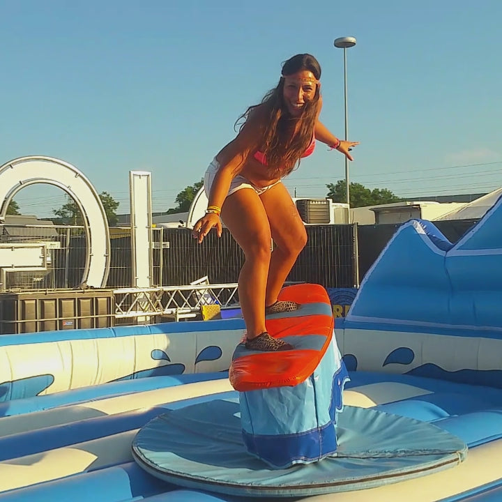 Eine Frau mit langen Haaren und ein Junge mit einem Tuch auf einem aufblasbarem Surf-Simulator zum mieten.