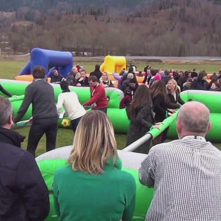 Teilnehmer sind an Stangen fixiert und bewegen sich wie bei einem Riesenwuzzler Tischfußballspiel, während sie versuchen, gemeinsam Tore zu erzielen – Teamwork und Spaß stehen im Mittelpunkt.